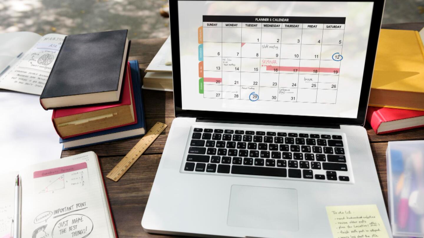 Laptop mit geöffnetem Kalender-Tool auf einem Holztisch, umgeben von Notizbüchern, Stiften und Linealen – Planungsumgebung für ein Event oder Projekt.