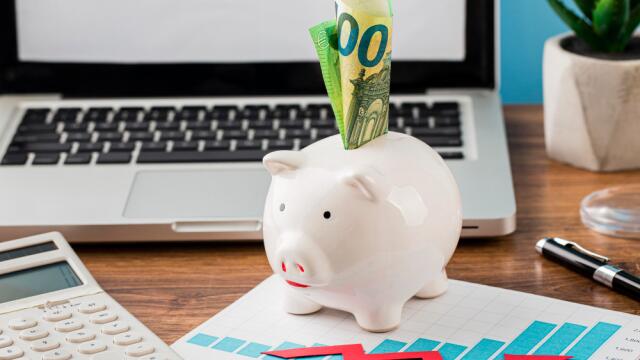 Ein weißes Sparschwein mit 100-Euro-Scheinen steht auf einem Schreibtisch vor einem Laptop. Daneben liegen ein Taschenrechner, ein Diagramm mit rotem Pfeil und eine Pflanze – Symbol für finanzielle Planung und Budgetoptimierung.