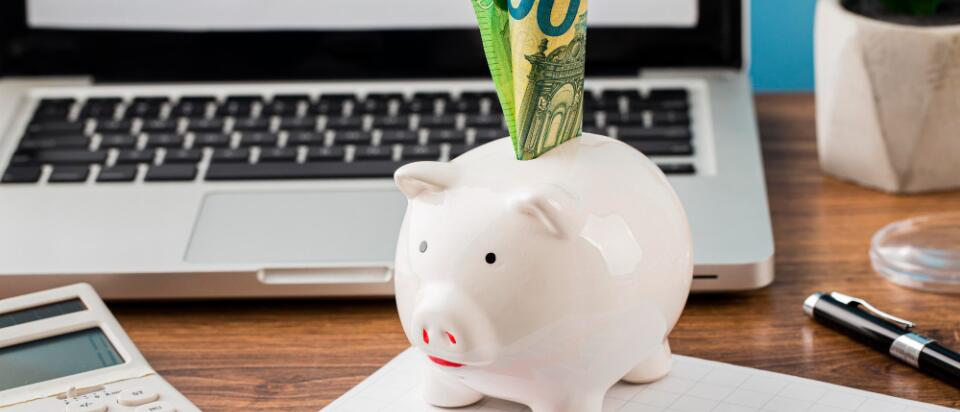 Ein weißes Sparschwein mit 100-Euro-Scheinen steht auf einem Schreibtisch vor einem Laptop. Daneben liegen ein Taschenrechner, ein Diagramm mit rotem Pfeil und eine Pflanze – Symbol für finanzielle Planung und Budgetoptimierung.