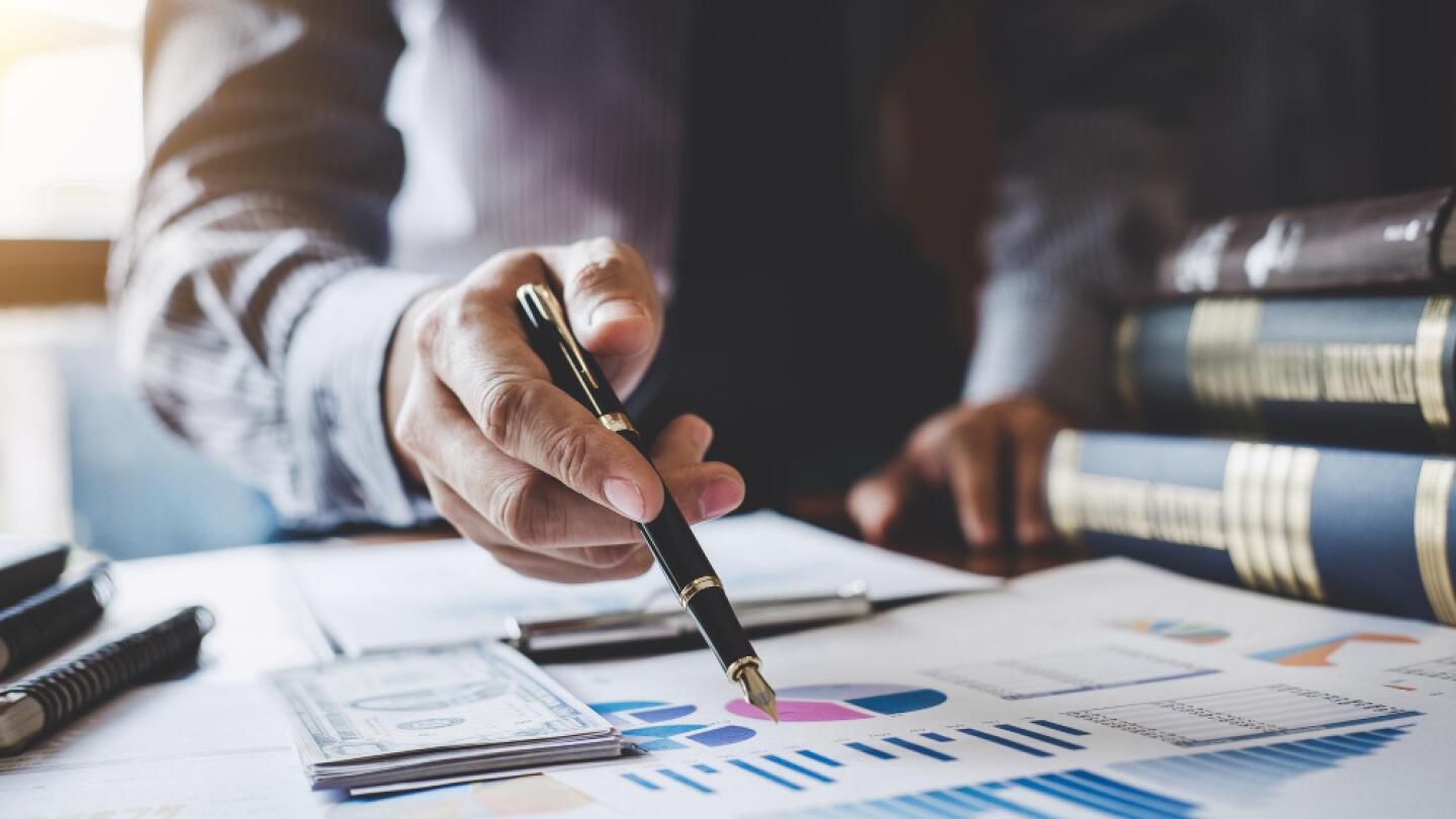 Geschäftsperson analysiert Finanzdiagramme und Budgetunterlagen am Schreibtisch, mit Geldbündeln, Diagrammen und Gesetzesbüchern im Hintergrund.