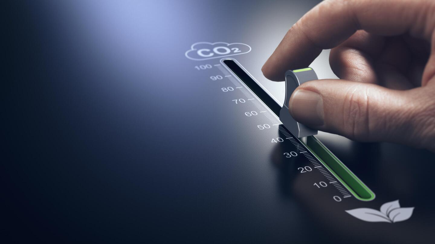 Eine Hand bewegt einen Schieberegler auf einer CO₂-Skala von einem hohen Wert in Richtung Null, was die Reduzierung von Emissionen symbolisiert.