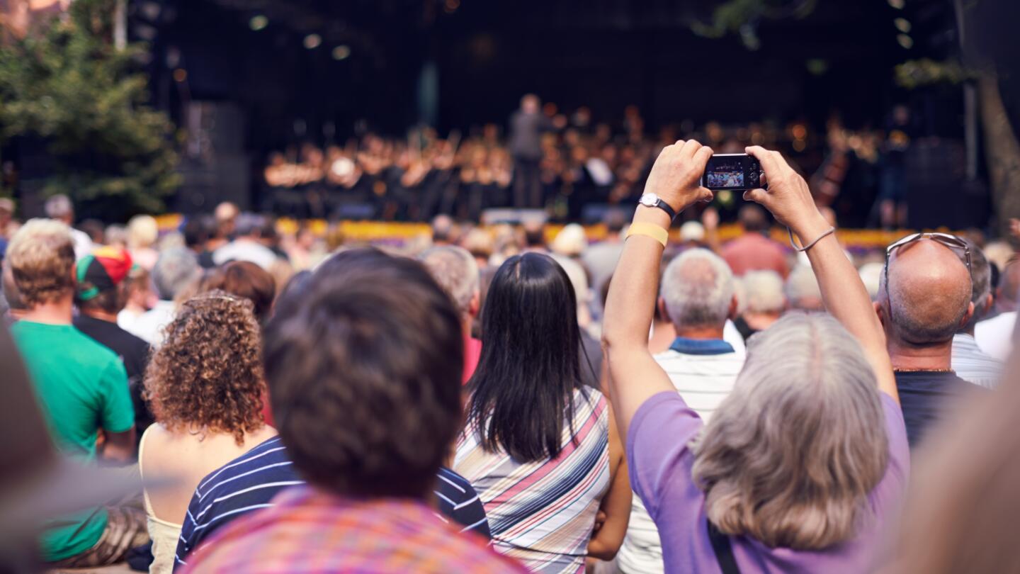 Zuschauermenge bei einem Outdoor-Event, wobei eine Person ein Smartphone hochhält, um ein Foto zu machen.