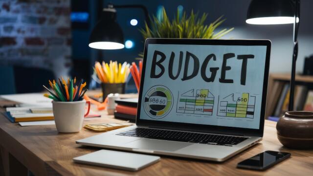 Ein Laptop steht auf einem Holzschreibtisch. Auf dem Bildschirm sind das Wort “BUDGET” und verschiedene Finanzdiagramme zu sehen. Die Szene wird von Schreibtischlampen beleuchtet und symbolisiert die fokussierte Finanz- und Event-Budgetplanung.