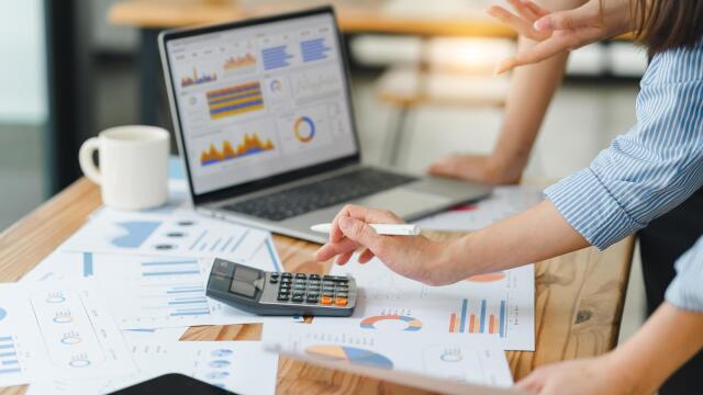 Nahaufnahme von Händen, die einen Taschenrechner auf einem Schreibtisch voller Finanzdiagramme und einem Laptop mit Datenvisualisierungen nutzen.