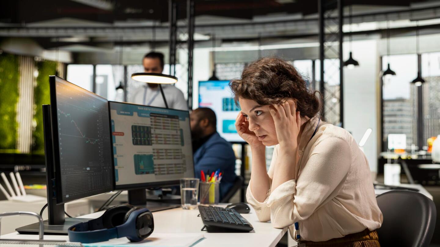 Eine konzentrierte, leicht gestresste Frau im modernen Büro vor mehreren Monitoren mit Daten-Dashboards; symbolisiert die Komplexität manueller Budgetsteuerung.