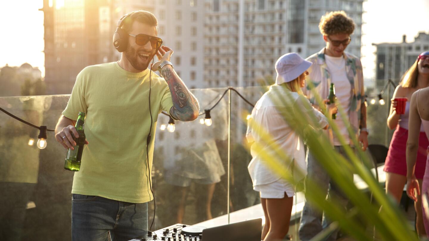 Ein DJ mit Kopfhörern legt bei einer Rooftop-Party im Sonnenuntergang auf, während Freunde um ihn herum tanzen und sich unterhalten.