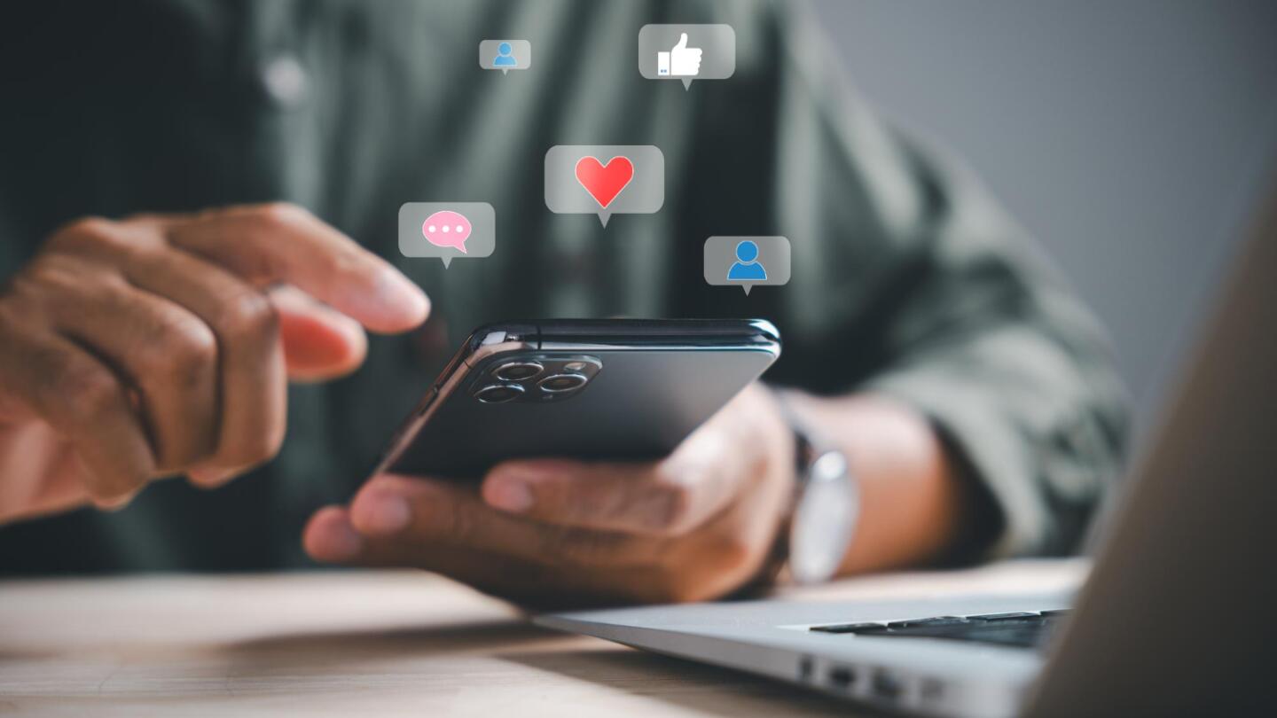 Hände halten ein Smartphone, über dem Social Media Symbole wie Likes und Herzen schweben.