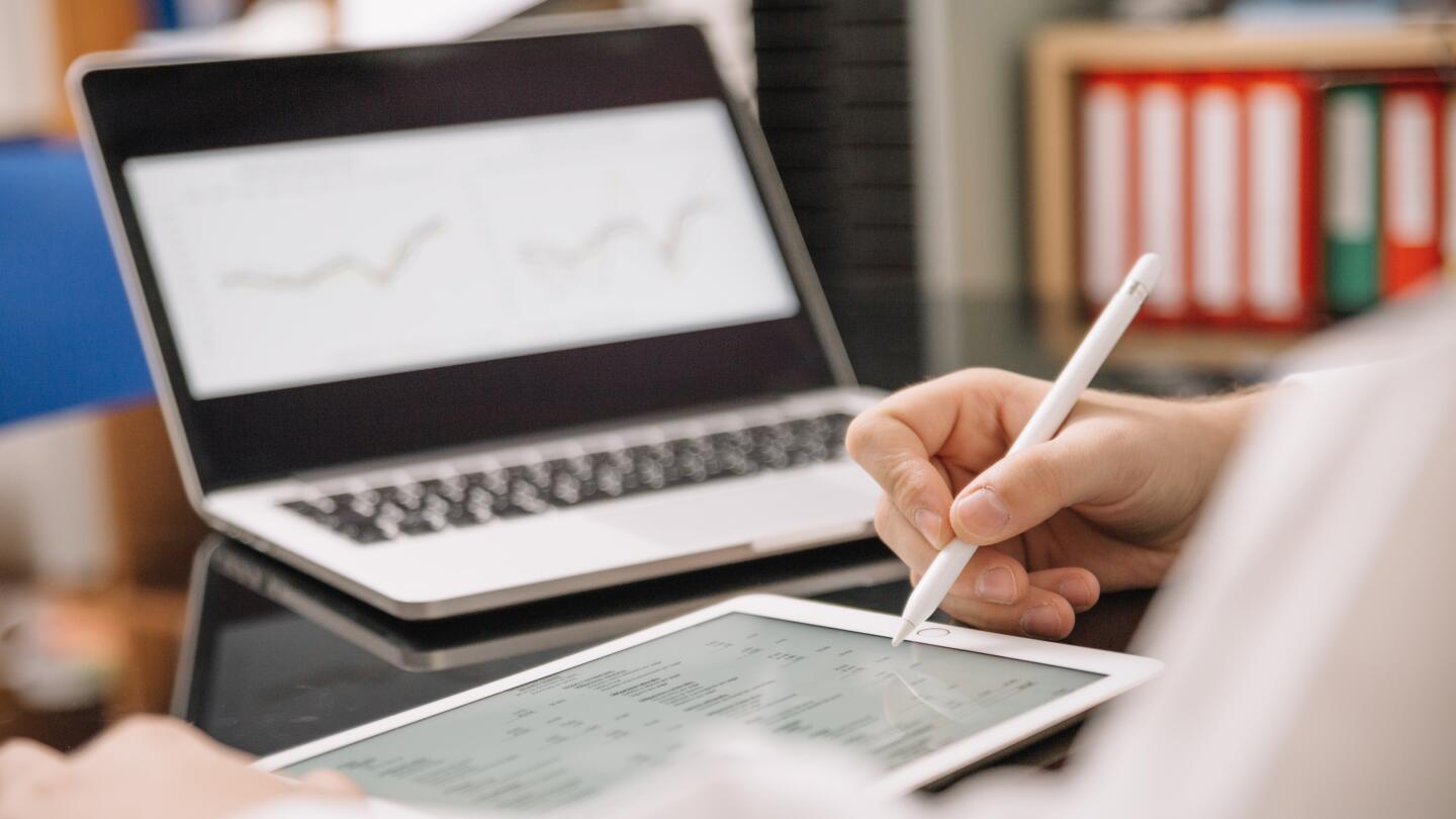 Im Hintergrund ist verschwommen ein Laptop mit Statistiken zu sehen. Der Fokus ist im Vordergrund, dort ist eine Hand zu sehen, die mit einem Stift auf einem Tablet arbeitet.