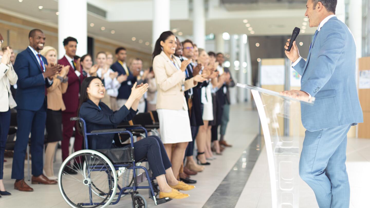 Ein vielfältiges Fachpublikum, darunter eine Frau im Rollstuhl, applaudiert stehend einem Redner auf einer Konferenz.
