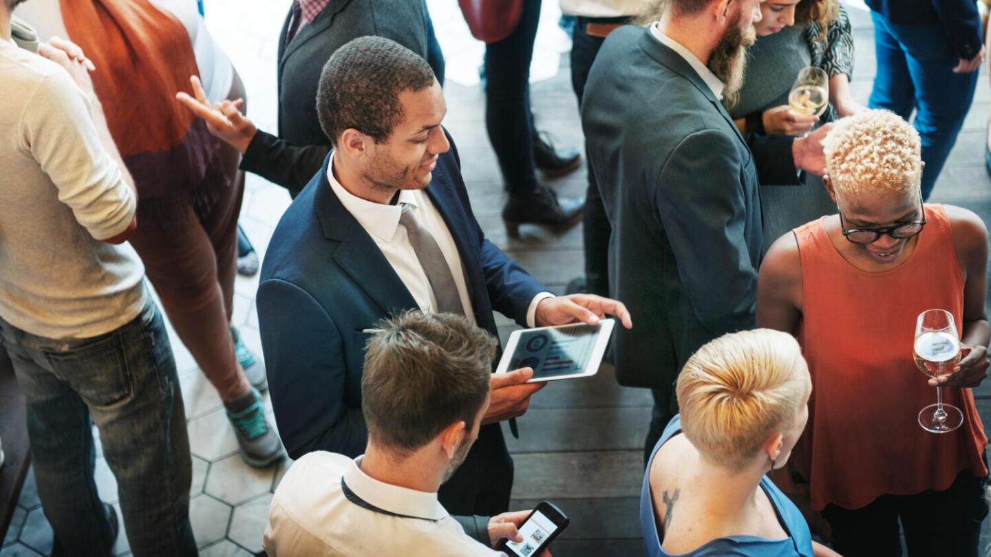 enschen in Business-Kleidung unterhalten sich bei einem Networking-Event in entspannter Atmosphäre. Einige halten Tablets oder Smartphones, andere ein Glas Wein. Die Szene vermittelt einen professionellen, aber lockeren Austausch unter Veranstaltungsteilnehmenden.