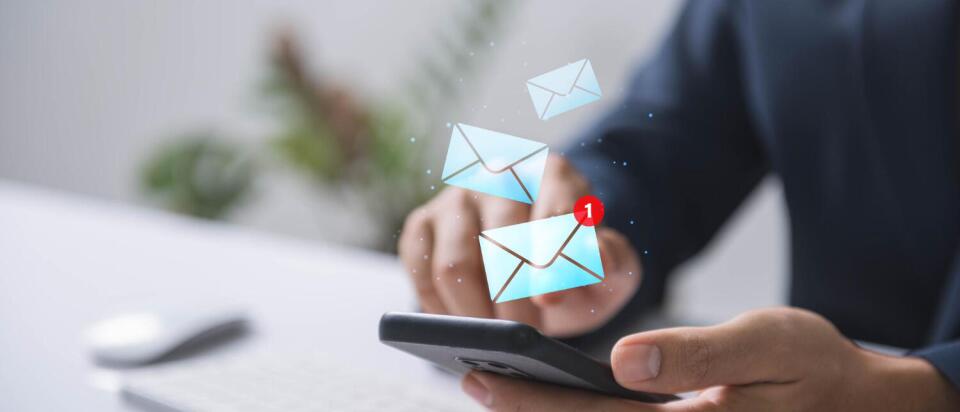 Person hält ein Smartphone in der Hand, auf dem eine neue E-Mail-Benachrichtigung angezeigt wird; im Hintergrund ist eine Computertastatur zu sehen.