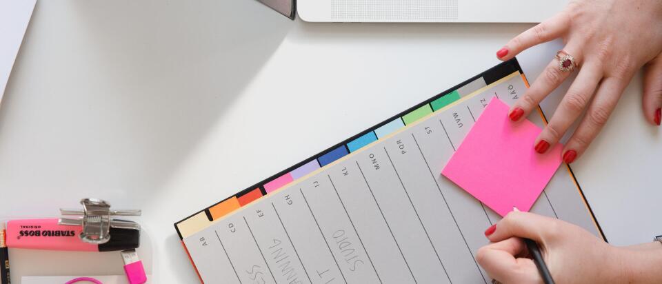 Eine Person sitzt an einem Schreibtisch mit einem Laptop, schreibt auf einen pinkfarbenen Notizzettel und hat eine Planungsmappe mit verschiedenen farbigen Registerkarten vor sich. Auf dem Tisch liegen außerdem Büroartikel wie ein Textmarker, Kopfhörer und ein Stift.
