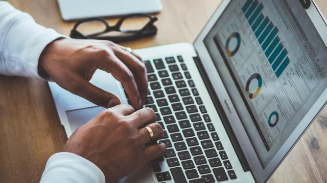 Eine Person arbeitet an einem Laptop, auf dessen Bildschirm Diagramme und Balkengrafiken zu sehen sind. Auf dem Tisch liegen außerdem eine Brille und ein Smartphone.