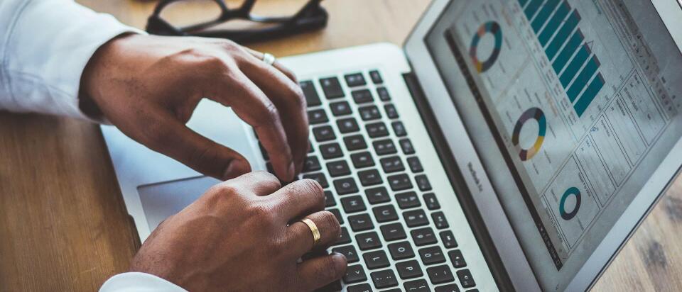 Eine Person arbeitet an einem Laptop, auf dessen Bildschirm Diagramme und Balkengrafiken zu sehen sind. Auf dem Tisch liegen außerdem eine Brille und ein Smartphone.