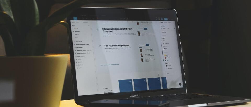 Ein Laptop steht auf einem kleinen Tisch neben einer Pflanze. Auf dem Bildschirm sind Webseiten oder Online-Dokumente mit Texten und Profilbildern zu sehen. Vor dem Laptop liegt ein Smartphone.