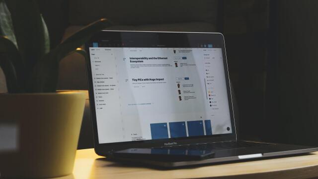 Ein Laptop steht auf einem kleinen Tisch neben einer Pflanze. Auf dem Bildschirm sind Webseiten oder Online-Dokumente mit Texten und Profilbildern zu sehen. Vor dem Laptop liegt ein Smartphone.