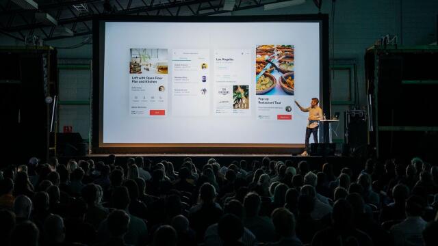 Ein Sprecher steht auf einer Bühne und präsentiert vor einem großen Publikum eine Bildschirmpräsentation mit verschiedenen App- oder Website-Designs, die auf einer großen Leinwand angezeigt werden.